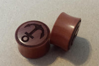 Load image into Gallery viewer, Two round, red-brown Flesh Plug Sawoholz Anker Seil Ring plugs with engraved anchor and rope designs rest on a light surface: one upright shows the motif clearly, while the other lies on its side. Ideal for an organic ear stretching style.
