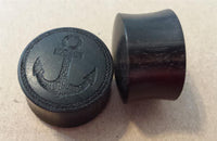 Load image into Gallery viewer, Two black circular plugs, one flat with an engraved anchor and rope design, rest on a plain light brown surface. These Flesh Plug Ebenholz Anker Seil Ring double flared Lobe Dehner schwarz Gravur offer an elegant organic piercing style.
