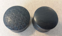 Load image into Gallery viewer, Two round black buttons on a beige surface; the left button resembles the Flesh Plug Ebenholz Stern Blume des Lebens with a black engraved geometric pattern, while the right button is smooth and plain.
