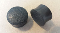 Load image into Gallery viewer, Two black Flesh Plug Ebenholz Stern Blume des Lebens double flared lobe plugs are shown, one displaying a geometric engraved pattern on top, the other revealing its smooth, curved side.
