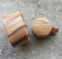 Load image into Gallery viewer, An Olivenholz Flesh Plug Lobe Piercing Holz Double Flared and its round lid with a small knob, both showcasing natural olive wood grain, sit atop a gray, speckled stone surface.
