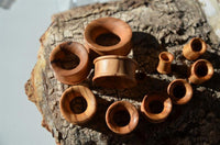 Load image into Gallery viewer, Various sizes of Olivenholz Flesh ribbed Tunnel Lobe Piercing Holz straight plugs are arranged on rustic bark, with sunlight highlighting the smooth, hand-carved wood and textured surfaces.
