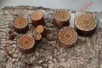 Load image into Gallery viewer, Plug Holz Tamarinde Krokodilholz in light and dark tones, each engraved with a Mandala Blüte design, are displayed on textured tree bark.
