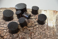 Load image into Gallery viewer, Several black cylindrical Flesh Plug Ebenholz Peace Knoten Muster double flared Lobe Dehner with engraved tops are arranged on tree bark, their glossy finish contrasting with the bark's rough, natural texture.
