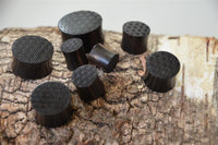 Load image into Gallery viewer, Several black cylindrical Flesh Plug Ebenholz Peace Knoten Muster double flared Lobe Dehner with engraved geometric patterns on top are arranged on rough tree bark, varying in size and featuring textured flat surfaces.
