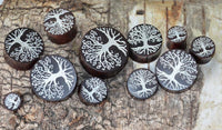 Load image into Gallery viewer, A set of Holz Plug Baum des Lebens Yggdrasil silber double flared plugs, crafted from wood and engraved with intricate white tree designs, displayed on a bark-textured surface. The plugs vary in size and feature detailed branching patterns.
