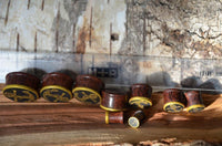 Load image into Gallery viewer, A set of Plug Holz Tamarinde dunkelbraun earplugs with golden brass-rimmed anchor and rope designs is arranged in a row on a wooden surface, with a transparent ruler and wood texture in the background.
