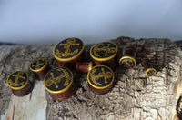 Load image into Gallery viewer, A set of Plug Holz Tamarinde dunkelbraun plugs with brass anchor and rope design, golden rim, and polished finish is displayed on textured bark; the set varies in size for piercing.

