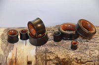 Load image into Gallery viewer, Holz Tunnel Eisenholz Rotholz schwarz braun, handgeschnitzt, are shown on a tree log. These wooden ear gauges feature smooth dark exteriors with lighter brown rims, highlighting their unique craftsmanship and style.
