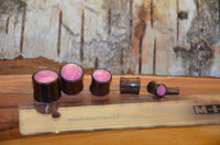 Load image into Gallery viewer, Five Holz Plug Sonoholz rosa schillernd Resin Inlay double flared plugs are lined up by size on a wood surface next to a clear ruler, in front of tree bark. Each features a shimmering pink resin center, ranging from large to small.

