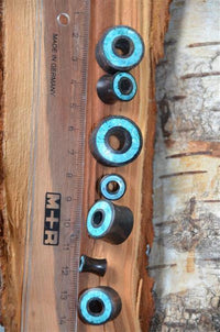 Load image into Gallery viewer, A clear M+R ruler marked Made in Germany lies on wood beside seven Flesh Tunnel Eisenholz schwarz Rim Crushed Türkis Inlay, arranged largest to smallest. A tree trunk appears in the background.

