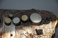 Load image into Gallery viewer, A set of Holz Plug Perlmutt Inlay Gravur Blume des Lebens ribbed, circular wooden containers with inlaid lids, arranged on a rough, sunlit tree bark surface.
