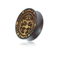 Load image into Gallery viewer, The Plug Holz hell dunkel Tribal Maske Gravur double flared is a round tamarind wood plug with an engraved tribal mask and decorative patterns, shown against a white background.
