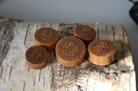 Load image into Gallery viewer, Five Holz Teakholz Plug Sonnen Maske double flared plugs, each featuring intricate engravings and varying sizes, are displayed on birch bark in a semi-circle, echoing the craftsmanship of Sonnen Krieger.
