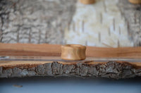 Load image into Gallery viewer, The Olivenholz Flesh Tunnel double flared abgerundet Holz stands upright on textured bark, with a blurred natural wood background, highlighting its handgeschnitzte, double-flared craftsmanship and light-toned finish.
