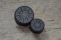 Load image into Gallery viewer, Two round Holz Flesh Plug Tamarinde Holz Krokodilholz Kreuz Ranken stamps with hand-carved, light floral patterns are set on rustic wood. One is significantly larger than the other.
