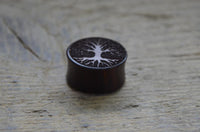 Load image into Gallery viewer, A Flesh Plug Holz Sonoholz Baum des Lebens double flared with a carved tree design on top sits on a light, textured wooden surface.
