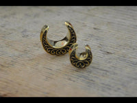 Load image into Gallery viewer, Two crescent-shaped Saddle Plug Messing antik gold Sichel Bögen Tunnel rings with black ornate patterns rest on a light wooden surface, showcasing intricate designs and a striking, elegant presence.
