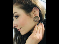 Load image into Gallery viewer, A woman with light skin and brown hair touches her ear, displaying a large, round Holz Flesh Plug Tamarinde Holz Krokodilholz Kreuz Ranken with an intricate swirling design. She has a nose piercing and long, dark nails.
