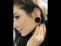 Load image into Gallery viewer, A woman with brown hair and light skin smiles slightly, showing off a large Flesh Plug Holz Parasitholz braun double flared ear gauge and a small nose stud. Her hand near her ear displays long, painted fingernails.
