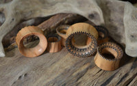 Load image into Gallery viewer, Handmade Holz Plug Olivenholz Sonnenblume double flared plugs with detailed carvings are displayed on a rustic wooden surface, accented by driftwood in the background.
