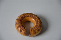 Load image into Gallery viewer, The Ohr Donut Piercing Pfau Mandala Jackfrucht Holz is a circular wooden bangle with a small opening, adorned with detailed dark brown mandala patterns, shown on a plain light gray surface.
