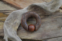 Load image into Gallery viewer, The Captive Bead Klemmkugel Ring Sawoholz Piercing Mandala, featuring intricate carved patterns and a smooth wooden bead, is displayed on driftwood with a rustic wooden background.
